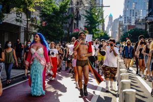 Rohan Zhou-Lee, a Black Asian nonbinary person in gray spandex shorts, leather harness, and red short cape, leads a diverse group of people in a march while holding a microphone.