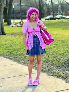 a person with magenta colored hair wearing a pink top and blue denim skirt with hot pink tote bag and shoes; they are standing on a sidewalk near a stretch of green grass