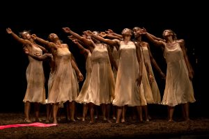 A line of several Black women dancers, wearing pale slip dresses and standing barefoot in earth. There is a strip of red cloth to the left on the ground. They are gesturing with their right arms to the left, their heads slightly bent.